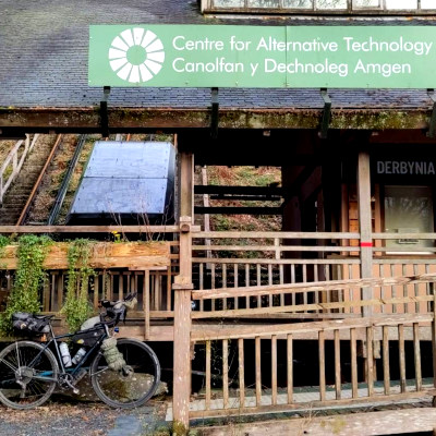 Wooden building front with tiled roof and Centre for Alternative Technology sign. In front is parked a well-laden bicycle.